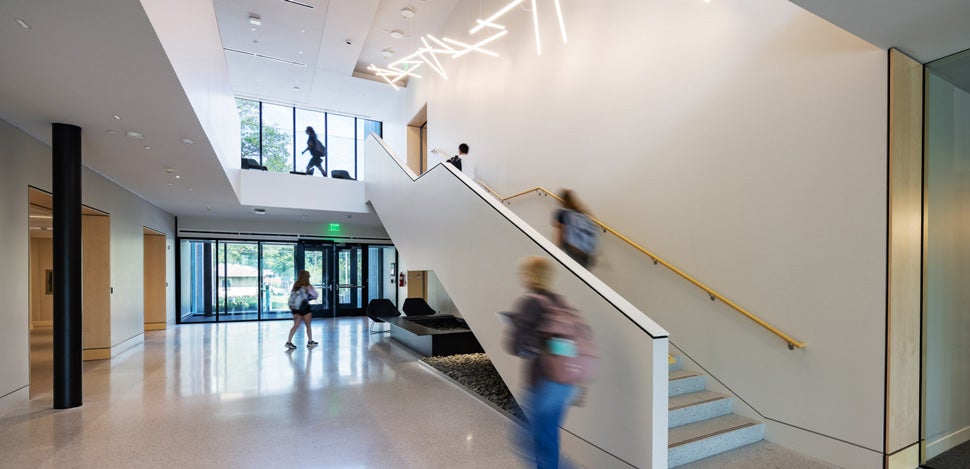 An atrium showing a staircase with students going up and down, floor-to-ceiling windows, and a modern light fixture design that spans the length of the staircase 