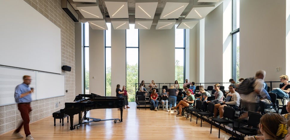 A choir room with strips of floor-to-ceiling windows, students in their seats, and a teacher at the front near a piano